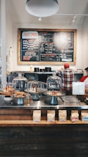 A cozy coffee shop interior with a large menu board on the wall, offering various drinks and food items. Two people are behind the counter, one wearing a red cap. On the counter, there are glass domes covering pastries, and bags of coffee are neatly arranged. The ambiance has a rustic charm with industrial lighting and wooden accents.