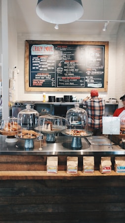 A cozy coffee shop interior with a large menu board on the wall, offering various drinks and food items. Two people are behind the counter, one wearing a red cap. On the counter, there are glass domes covering pastries, and bags of coffee are neatly arranged. The ambiance has a rustic charm with industrial lighting and wooden accents.