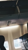 The interior of an airplane cabin featuring empty passenger seats with dark upholstery and partially open overhead compartments. A small section of the aircraft window is visible, allowing soft natural light to fill the space. The ambiance is quiet and calm with no passengers present.