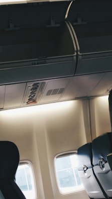 The interior of an airplane cabin featuring empty passenger seats with dark upholstery and partially open overhead compartments. A small section of the aircraft window is visible, allowing soft natural light to fill the space. The ambiance is quiet and calm with no passengers present.