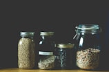 Four glass jars contain various grains and seeds, neatly arranged in a row. The jars vary in size, with metal lids. The contents include tri-color quinoa, black beans, and a mix of seeds and grains, all displayed against a dark background.