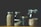 Four glass jars contain various grains and seeds, neatly arranged in a row. The jars vary in size, with metal lids. The contents include tri-color quinoa, black beans, and a mix of seeds and grains, all displayed against a dark background.