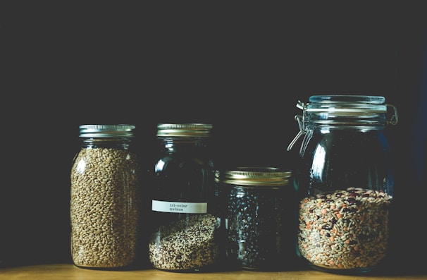 Four glass jars contain various grains and seeds, neatly arranged in a row. The jars vary in size, with metal lids. The contents include tri-color quinoa, black beans, and a mix of seeds and grains, all displayed against a dark background.