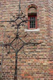 An ornate black wrought iron structure is mounted on a brick wall. The wall is made of various shades of brown and red bricks. There is a small arched window with a red frame and a grid of glass panes. A metal pipe runs vertically along the wall near the structure.