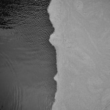 Aerial view of a distinct boundary between a textured, dark liquid surface and a lighter, granular solid surface. The separation line creates an irregular, flowing edge.