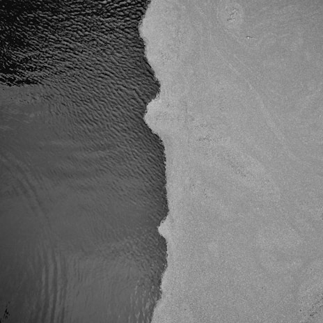 Aerial view of a distinct boundary between a textured, dark liquid surface and a lighter, granular solid surface. The separation line creates an irregular, flowing edge.