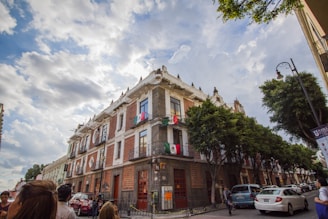 A large historical building with ornate architectural details is situated on a bustling street corner. The building is adorned with Mexican flags, and lush green trees line the sidewalk. The sky is partly cloudy, casting soft shadows on the scene. Several people and cars can be seen in the foreground, suggesting an urban environment.
