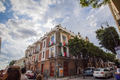 A large historical building with ornate architectural details is situated on a bustling street corner. The building is adorned with Mexican flags, and lush green trees line the sidewalk. The sky is partly cloudy, casting soft shadows on the scene. Several people and cars can be seen in the foreground, suggesting an urban environment.