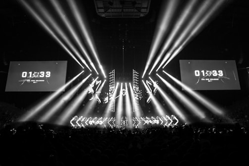 A black and white image captures a dramatic indoor scene featuring a large stage setup with intense beams of light radiating from the center. Two large screens are prominently displaying the text '01:33 ESL One Cologne 2017'. The audience can be seen seated in the dark foreground, while angular patterns can be spotted on the stage.