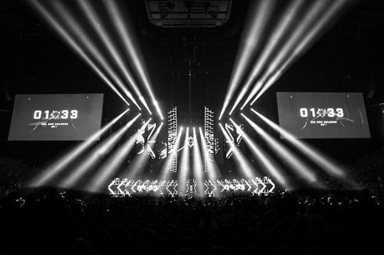 A black and white image captures a dramatic indoor scene featuring a large stage setup with intense beams of light radiating from the center. Two large screens are prominently displaying the text '01:33 ESL One Cologne 2017'. The audience can be seen seated in the dark foreground, while angular patterns can be spotted on the stage.