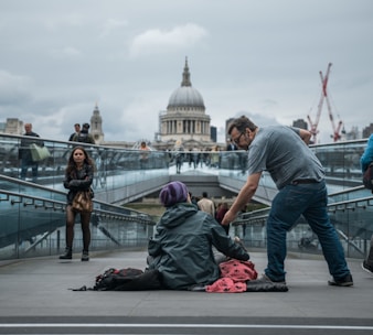 A man bends down to offer something to a person sitting on the ground, wrapped in a blanket and wearing a beanie. The scene is set on a modern pedestrian bridge with a view of a grand domed building in the background. Other pedestrians walk by, some paying attention while others are preoccupied.