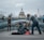 A man bends down to offer something to a person sitting on the ground, wrapped in a blanket and wearing a beanie. The scene is set on a modern pedestrian bridge with a view of a grand domed building in the background. Other pedestrians walk by, some paying attention while others are preoccupied.