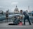 A man bends down to offer something to a person sitting on the ground, wrapped in a blanket and wearing a beanie. The scene is set on a modern pedestrian bridge with a view of a grand domed building in the background. Other pedestrians walk by, some paying attention while others are preoccupied.