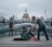 A man bends down to offer something to a person sitting on the ground, wrapped in a blanket and wearing a beanie. The scene is set on a modern pedestrian bridge with a view of a grand domed building in the background. Other pedestrians walk by, some paying attention while others are preoccupied.