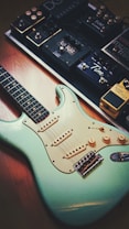 A turquoise electric guitar is placed on a wooden floor next to a row of various guitar effects pedals. The guitar has a classic design with three single-coil pickups, a white pickguard, and control knobs. The pedals are arranged on a pedalboard, featuring different brands and types, each with distinct knobs and foot switches.