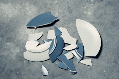 Several broken pieces of a ceramic plate are scattered on a rough, gray concrete surface. The fragments show variations in size and shape, with some pieces displaying both blue and white colors.