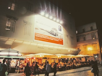An outdoor cafe at night with people dining under large umbrellas. The building behind features a large illuminated billboard advertising a car. Small kiosks or shops are seen to the left, contributing to a vibrant evening atmosphere.