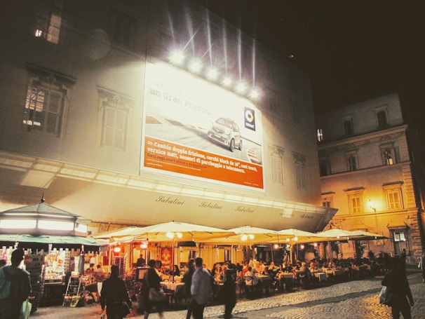 An outdoor cafe at night with people dining under large umbrellas. The building behind features a large illuminated billboard advertising a car. Small kiosks or shops are seen to the left, contributing to a vibrant evening atmosphere.