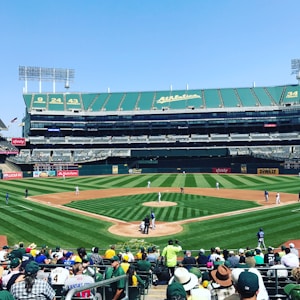A baseball game is underway in a large stadium. Several players are positioned around the field, with one team wearing white uniforms. The stadium seats are mostly empty except for a group of spectators sitting near the foreground. Green and yellow colors dominate the stadium's decor, and advertisements are visible on the field's periphery.