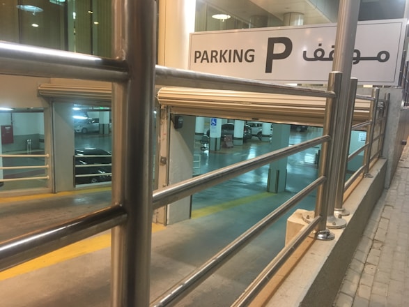 An indoor parking garage with multiple levels is visible. A prominent sign in both English and Arabic indicates parking directions. The scene includes metal railings, parked cars, and visible parking lane markers.