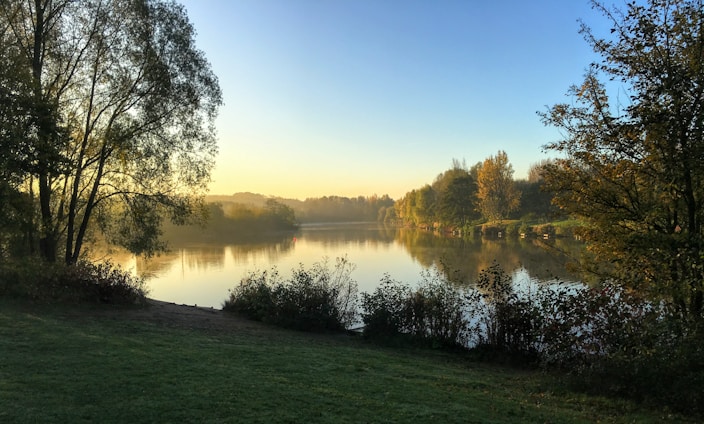 A serene morning scene with a person meditating beside a calm lake surrounded by soft greenery.