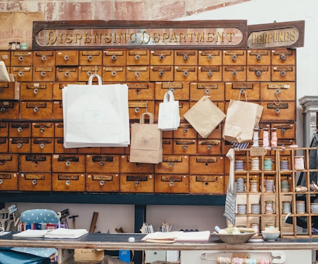 A vintage wooden cabinet with numerous small drawers is mounted on the wall. Signs reading 'Dispensing Department' and 'Perfumes' are displayed above the drawers. Various sizes of paper bags with handles hang from some of the drawers. Below, a counter holds ribbons, open books, and other small items.