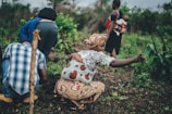 Several people are engaged in agricultural activities in a lush, green setting. A woman, with a baby strapped to her back, appears to be pointing or reaching out, while others around her tend to the plants.