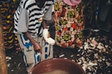 People are standing around a large plastic basin filled with water, preparing what appears to be food. The individuals are wearing brightly patterned clothing, and one person is holding a piece of food in their hand, possibly preparing or peeling it. The ground is covered with debris, indicating a food preparation area.