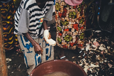 People are standing around a large plastic basin filled with water, preparing what appears to be food. The individuals are wearing brightly patterned clothing, and one person is holding a piece of food in their hand, possibly preparing or peeling it. The ground is covered with debris, indicating a food preparation area.