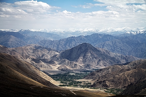 A vast landscape featuring rugged, arid mountains and valleys. Snow-capped peaks are visible in the background, while the foreground includes rocky terrain with some patches of greenery.