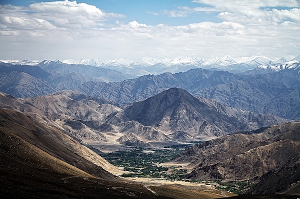 A vast landscape featuring rugged, arid mountains and valleys. Snow-capped peaks are visible in the background, while the foreground includes rocky terrain with some patches of greenery.