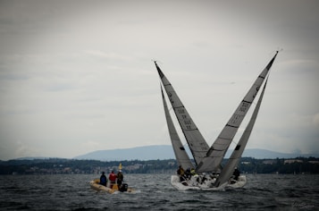 Two sailboats with tall sails navigate through the water, with a motorized boat nearby carrying several people. The scene takes place on a vast body of water with a distant shoreline lined with buildings and hills under a cloudy sky.