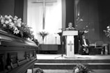 A man in religious robes stands at a podium with arms outstretched, speaking in a chapel. A large cross is prominently displayed on the wall behind him. A casket adorned with flowers is visible in the foreground to the left, and there are flower arrangements on either side. Another man in a suit is seated in the background.