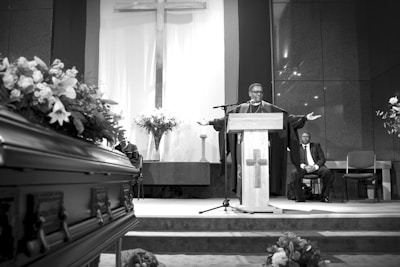 A man in religious robes stands at a podium with arms outstretched, speaking in a chapel. A large cross is prominently displayed on the wall behind him. A casket adorned with flowers is visible in the foreground to the left, and there are flower arrangements on either side. Another man in a suit is seated in the background.