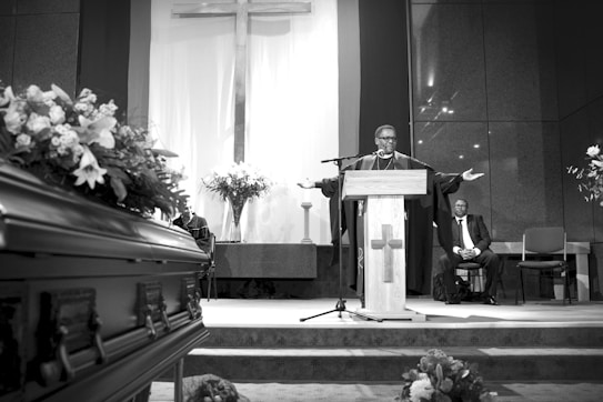 A man in religious robes stands at a podium with arms outstretched, speaking in a chapel. A large cross is prominently displayed on the wall behind him. A casket adorned with flowers is visible in the foreground to the left, and there are flower arrangements on either side. Another man in a suit is seated in the background.