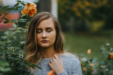 A young woman with closed eyes stands amidst greenery and flowers, gently touching her neck. She appears to be calm and serene, with a soft expression on her face. A single orange rose is prominently visible near her head.