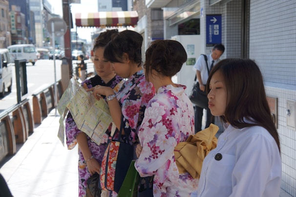 A group of women wearing colorful kimono stand on a city sidewalk, examining a map. The street is lined with buildings and a few vehicles are visible in the background. A man in white shirt appears in the background, leaning against a wall.