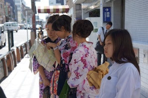 A group of women wearing colorful kimono stand on a city sidewalk, examining a map. The street is lined with buildings and a few vehicles are visible in the background. A man in white shirt appears in the background, leaning against a wall.