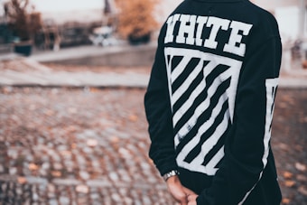A person is wearing a black sweatshirt with bold white text and a graphic design on the back, standing in an outdoor setting with cobblestone pavement and blurred background elements, possibly including trees and outdoor furniture.