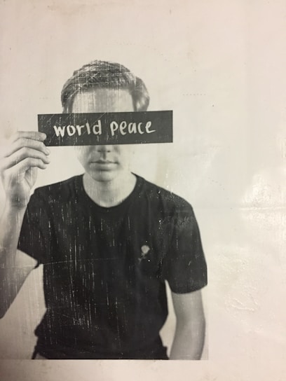 A person is holding a card with the words 'world peace' blocking their eyes, creating a thought-provoking and contemplative scene. The image is in black and white with a slightly textured appearance.