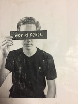 A person is holding a card with the words 'world peace' blocking their eyes, creating a thought-provoking and contemplative scene. The image is in black and white with a slightly textured appearance.