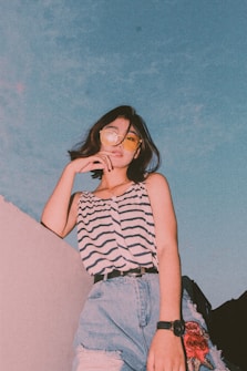 A person poses confidently, gazing forward with one hand resting on their chin. They wear a sleeveless striped top, denim shorts, and yellow-tinted sunglasses. The background features a vibrant blue sky, adding to the casual and relaxed vibe of the scene.