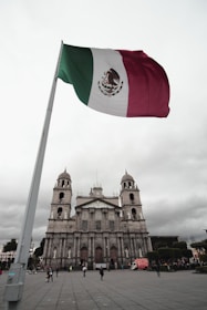 A large Mexican flag is prominently displayed against a cloudy sky, with a historical stone cathedral featuring two bell towers in the background. Several people are walking or standing in the plaza in front of the cathedral, and a red truck is parked nearby.