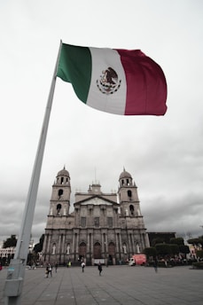 A large Mexican flag is prominently displayed against a cloudy sky, with a historical stone cathedral featuring two bell towers in the background. Several people are walking or standing in the plaza in front of the cathedral, and a red truck is parked nearby.