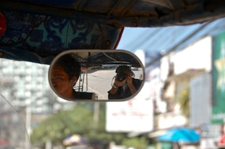 The rearview mirror of a vehicle shows two people, one driving and the other taking a photograph with a camera. The background includes an urban environment with blurred buildings and signs.