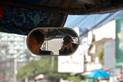 The rearview mirror of a vehicle shows two people, one driving and the other taking a photograph with a camera. The background includes an urban environment with blurred buildings and signs.