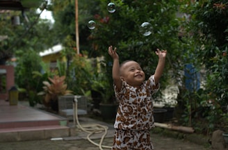 A young child stands outdoors, joyfully reaching up towards bubbles floating above. The child is wearing a patterned outfit and appears to be in a lush garden setting with greenery and potted plants around. The background includes a hose and a small building structure.