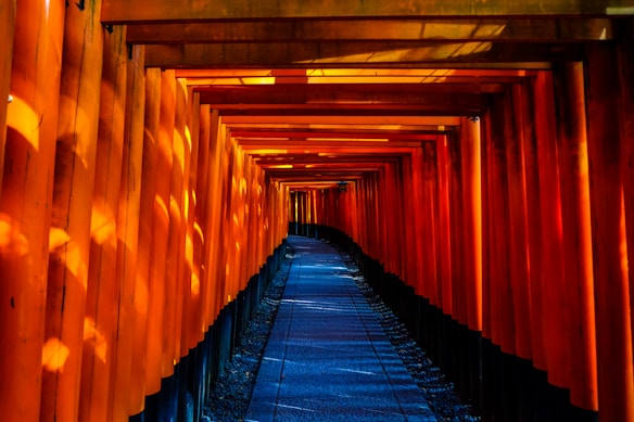 A pathway is surrounded by a series of bright orange torii gates creating a tunnel-like structure. The path is dimly lit with shadows of sunlight creating a rhythmic pattern on the gates. The perspective view leads to the distant end of the pathway, emphasizing depth and symmetry.