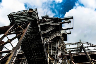 A complex industrial structure with intricate metal frameworks and pipes, set against a backdrop of a cloudy sky. The perspective is from below, emphasizing the scale and architectural intricacies of the structure.