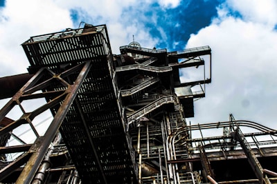 A complex industrial structure with intricate metal frameworks and pipes, set against a backdrop of a cloudy sky. The perspective is from below, emphasizing the scale and architectural intricacies of the structure.
